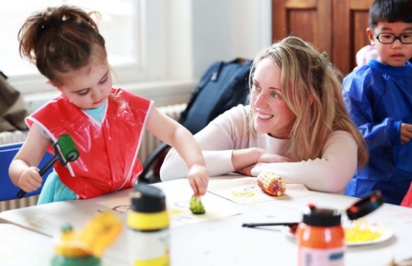 A child is painting wiyh a paint roller and sponge. An adult sits beside the child and is watching and smiling.