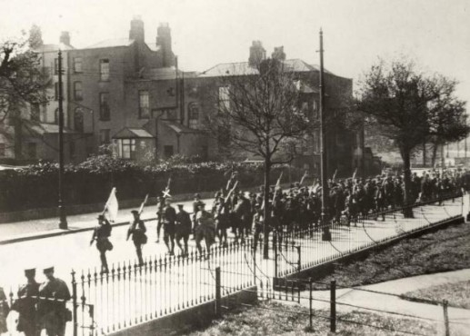 Surrender of Boland’s Mill Garrison. Men from the 3rd Battalion of the Irish Volunteers being led down Northumberland Road after the surrender of the Boland’s Mill Garrison carrying weapons and a flag. Eamonn De Valera is marked by the white X above his head. Credit: National Library of Ireland.