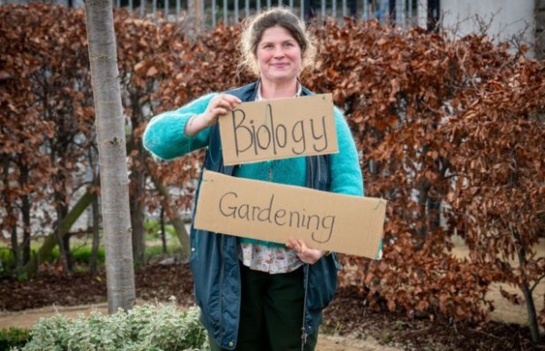 A person in a garden holding two cardboard signs, one reading ‘Biology’ and the other ‘Gardening’.