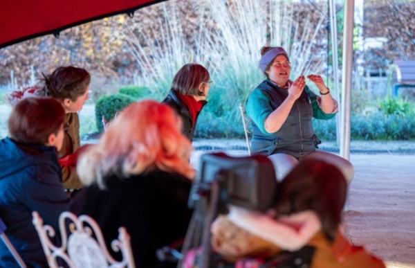 A group of people are in a semi circle under a tent, looking at a person demonstrating with their hands.