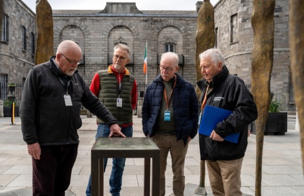 Four people standing in front of a building with an Irish flag. One person is pointing at a plinth. 