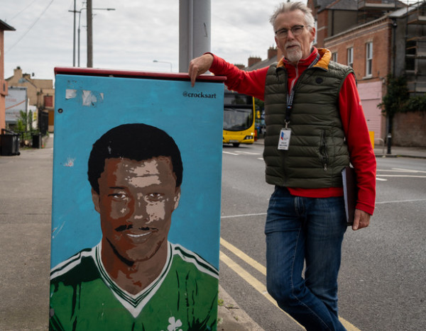 A man is standing on the roadside leaning on an artwork featuring a man in an Irish football jersey