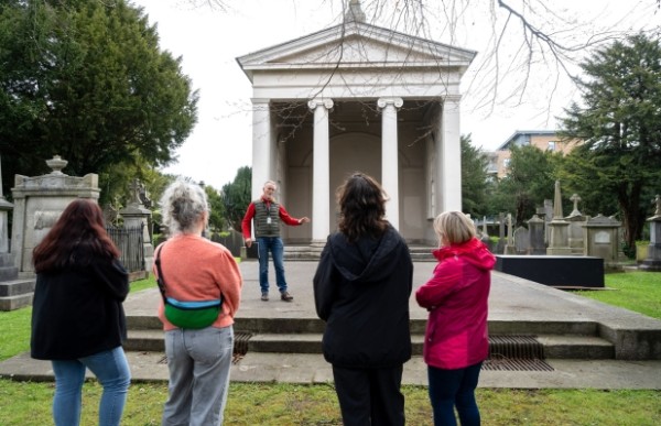 A man is standing in front of a large building in a graveyard, giving a talk to four people who have their backs to the camera