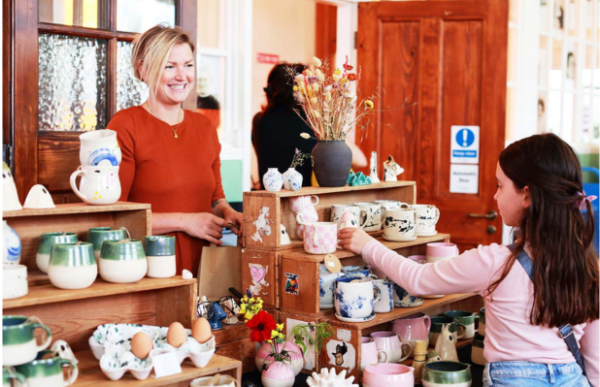 A woman smiles as she stands behind a stall displaying handmade pottery, while a young person browses the items on the shelves.