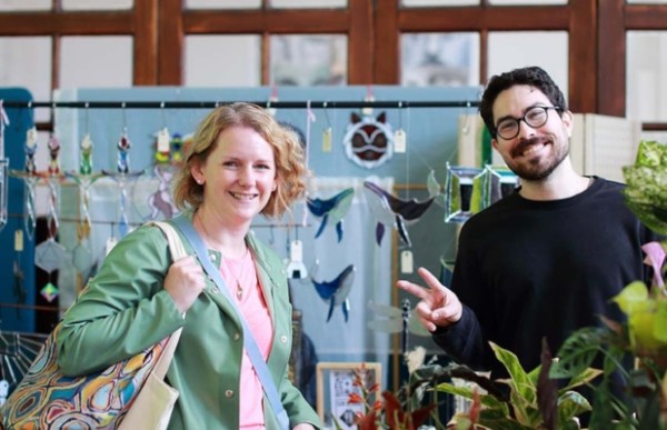 Two people smiling at the camera, standing beside plants and in front of a hanging display of stained glass.
