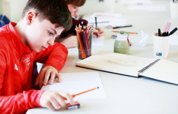 A child is sitting at a desk, drawing on a sketchpad with colouring pencils
