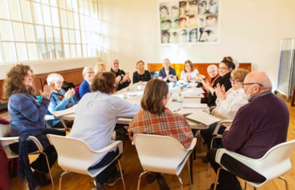 A group of adults are sitting at a large table. They have notebooks on the table, and they are clapping their hands. 